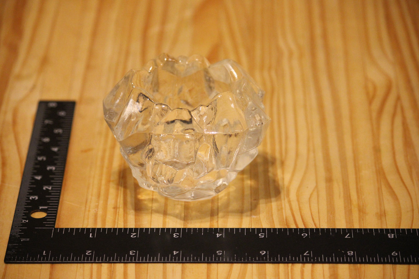 A clear faceted glass candle holder placed on a light wood surface next to a black ruler for scale.