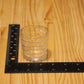 A clear glass votive candle holder with stacked ridges sits on a light wood surface next to a black L-square ruler showing measurements.
