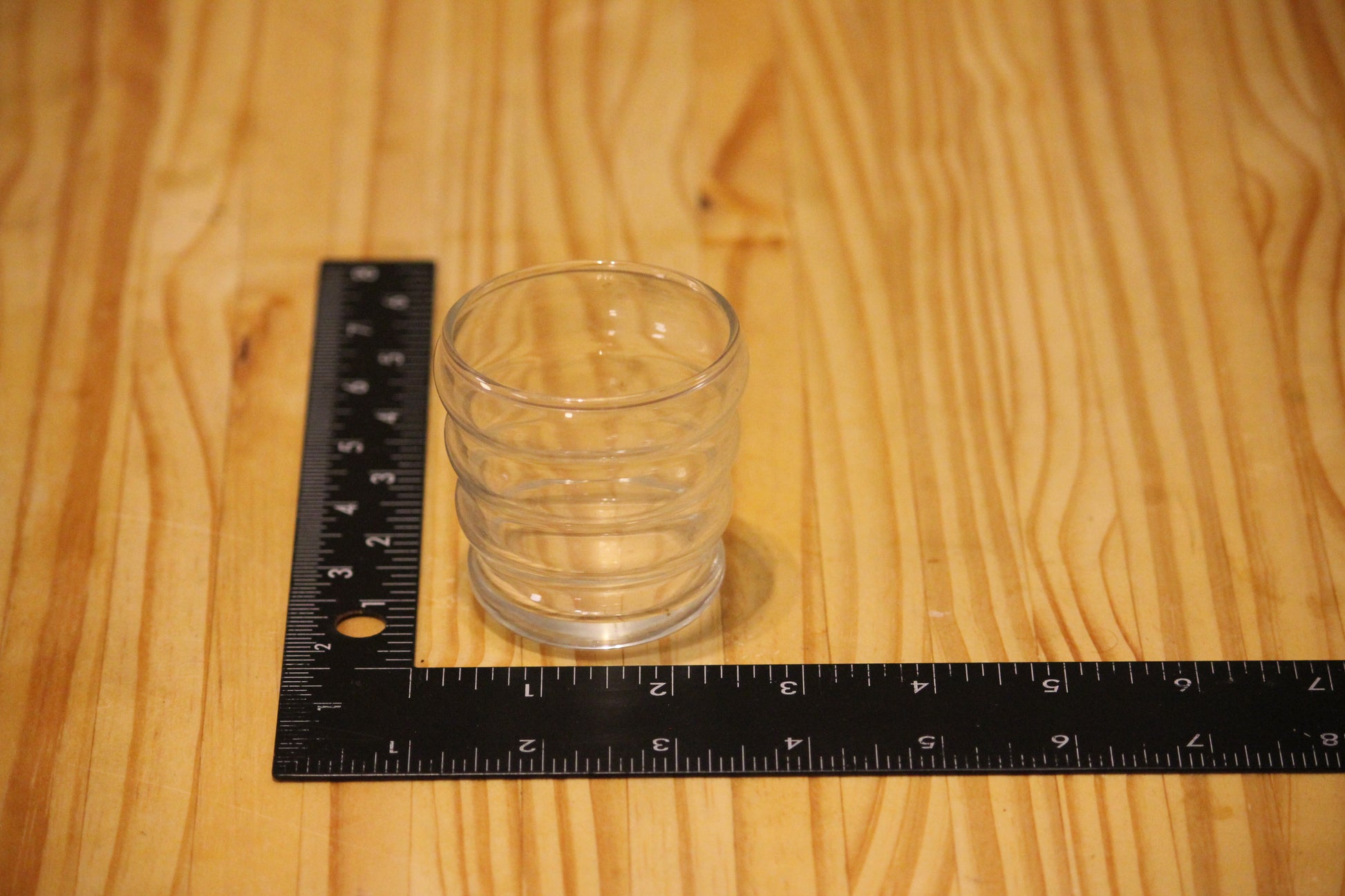 A clear glass votive candle holder with stacked ridges sits on a light wood surface next to a black L-square ruler showing measurements.
