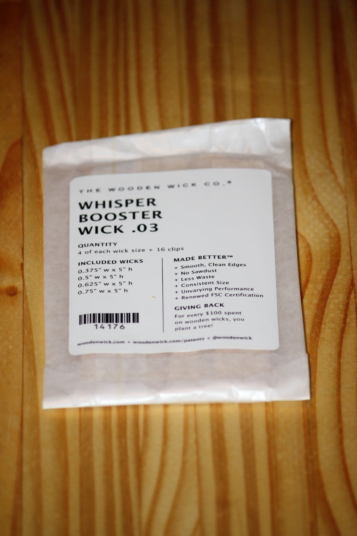 A small white packet of The Wooden Wick Co. Whisper Booster Wick .03, placed on a light brown wooden surface.