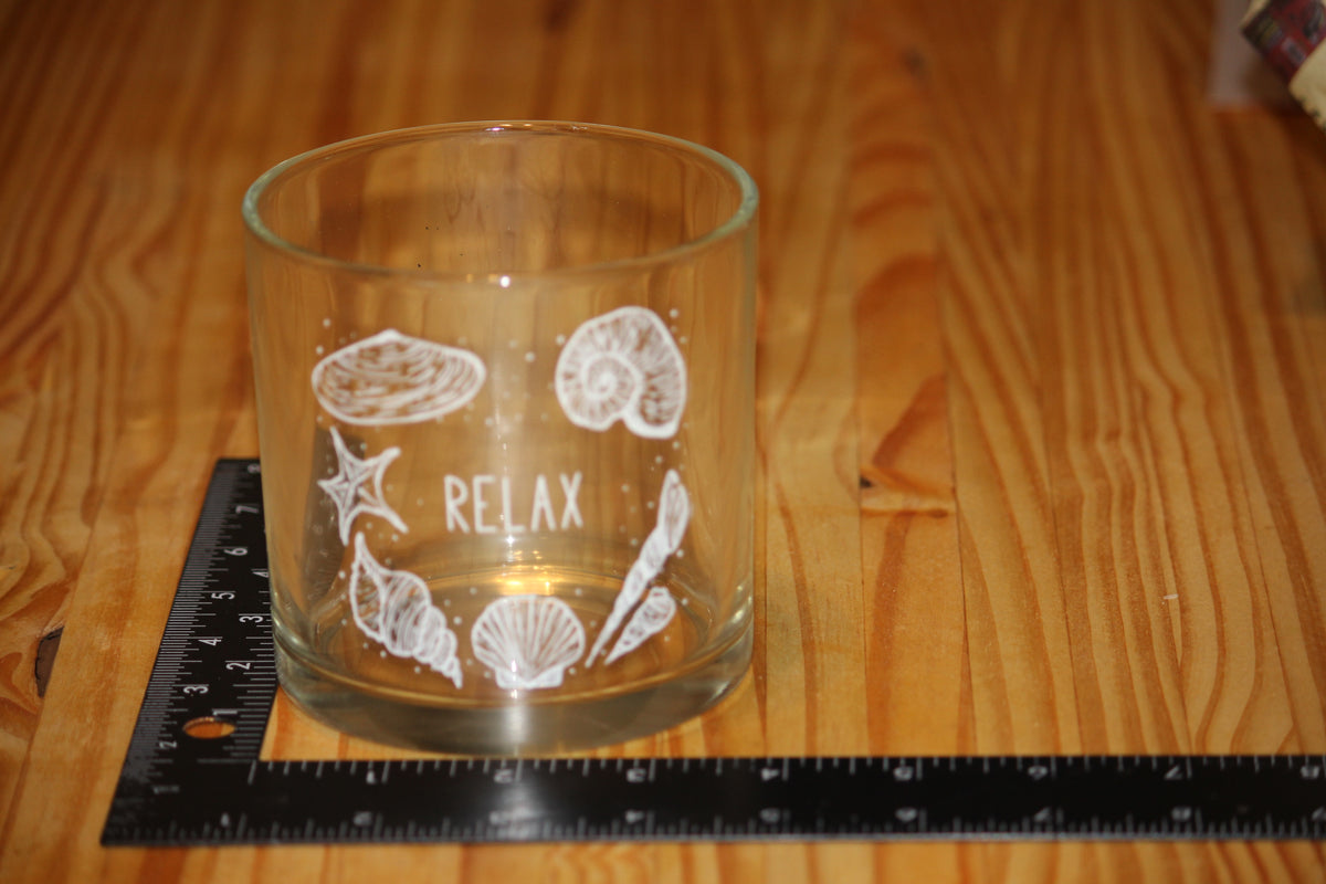 A clear glass candle holder with white sea-themed illustrations and the word 'RELAX' printed on it, placed on a wooden table next to a black ruler for scale.