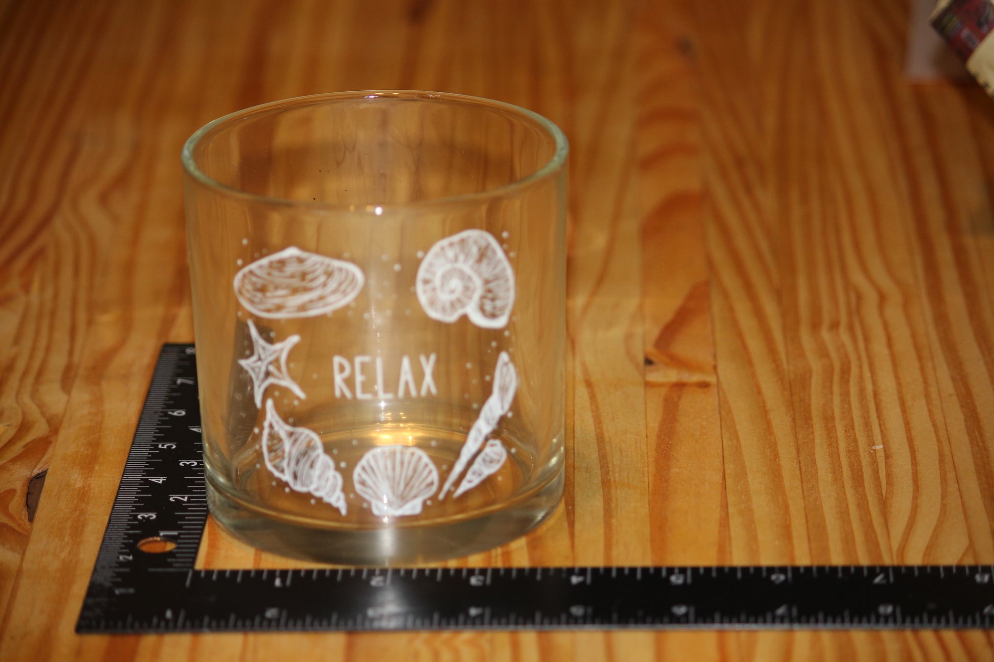 A clear glass candle holder with white sea-themed illustrations and the word 'RELAX' printed on it, placed on a wooden table next to a black ruler for scale.