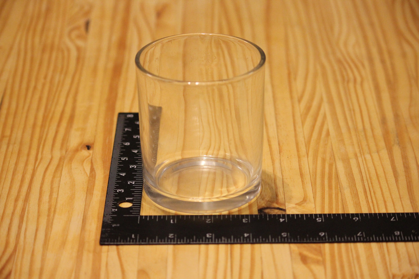 A clear glass candle holder placed on a black measuring square over a light wood grain surface, showing its dimensions.