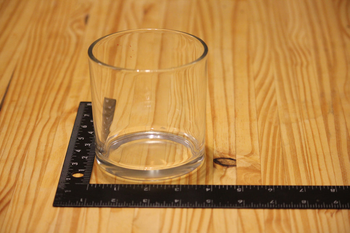 A clear glass candle holder with a thick base sits on a wooden table next to a black ruler, showing its height.