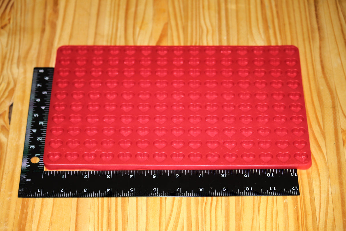A red rectangular silicone mold tray with heart cavities, shown on a light wood surface next to a black measuring ruler to indicate size.