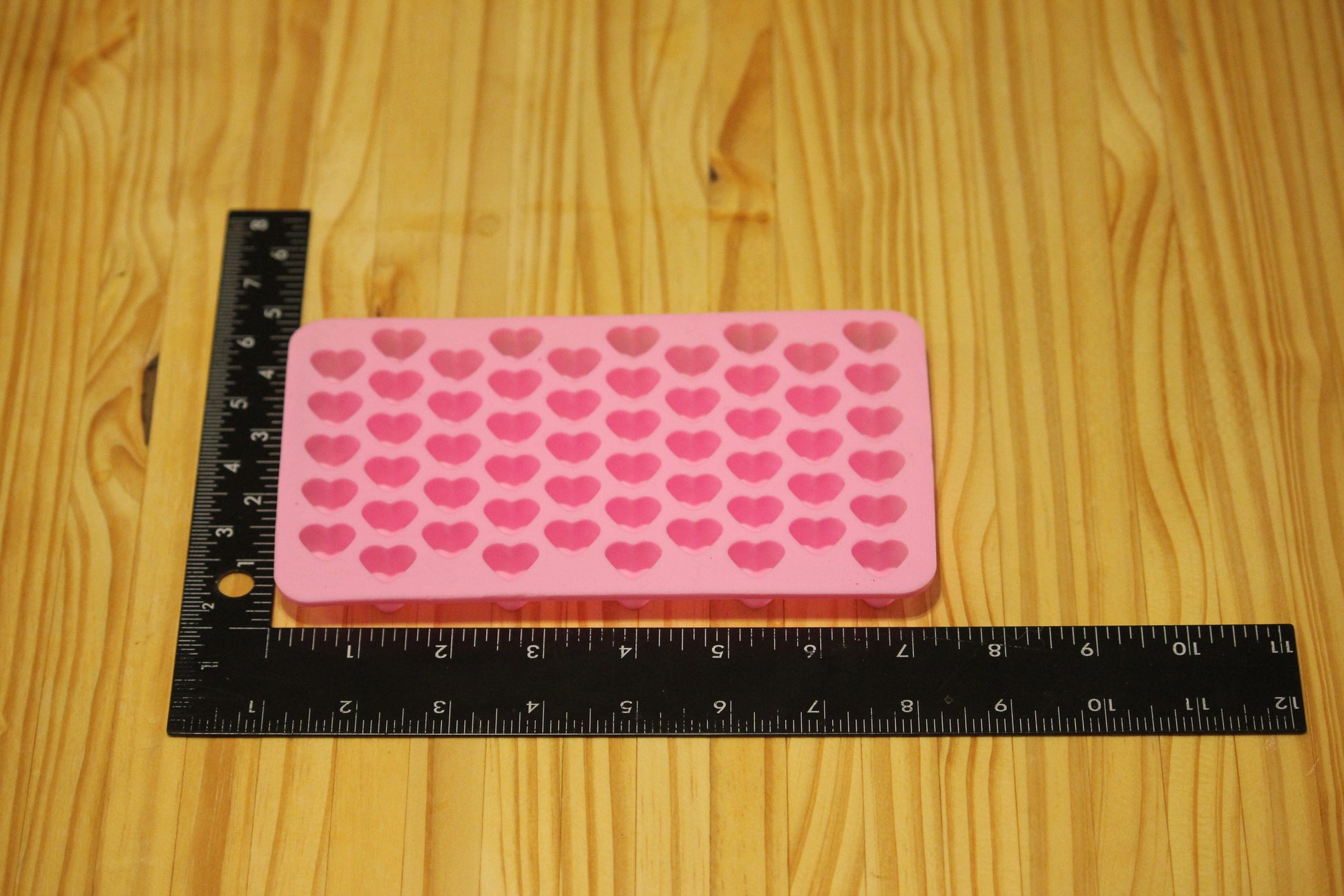 A pink silicone mold with rows of heart-shaped cavities, shown on a light wood surface next to a black ruler for scale.