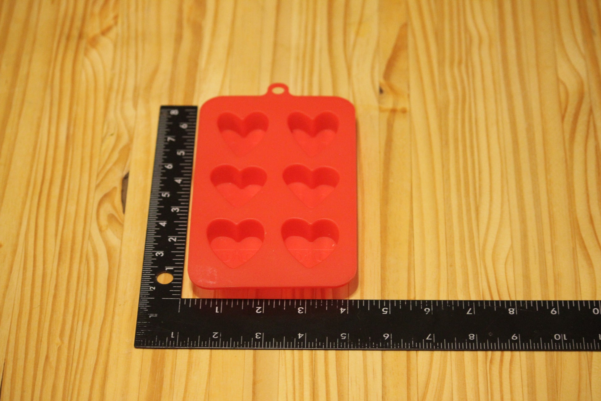 A red silicone mold with six heart-shaped cavities, shown on a light wood grain surface next to a black ruler for scale.