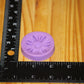 A small lavender purple circular mold with a flower embossed design, shown next to a black ruler on a wooden surface to indicate its size.