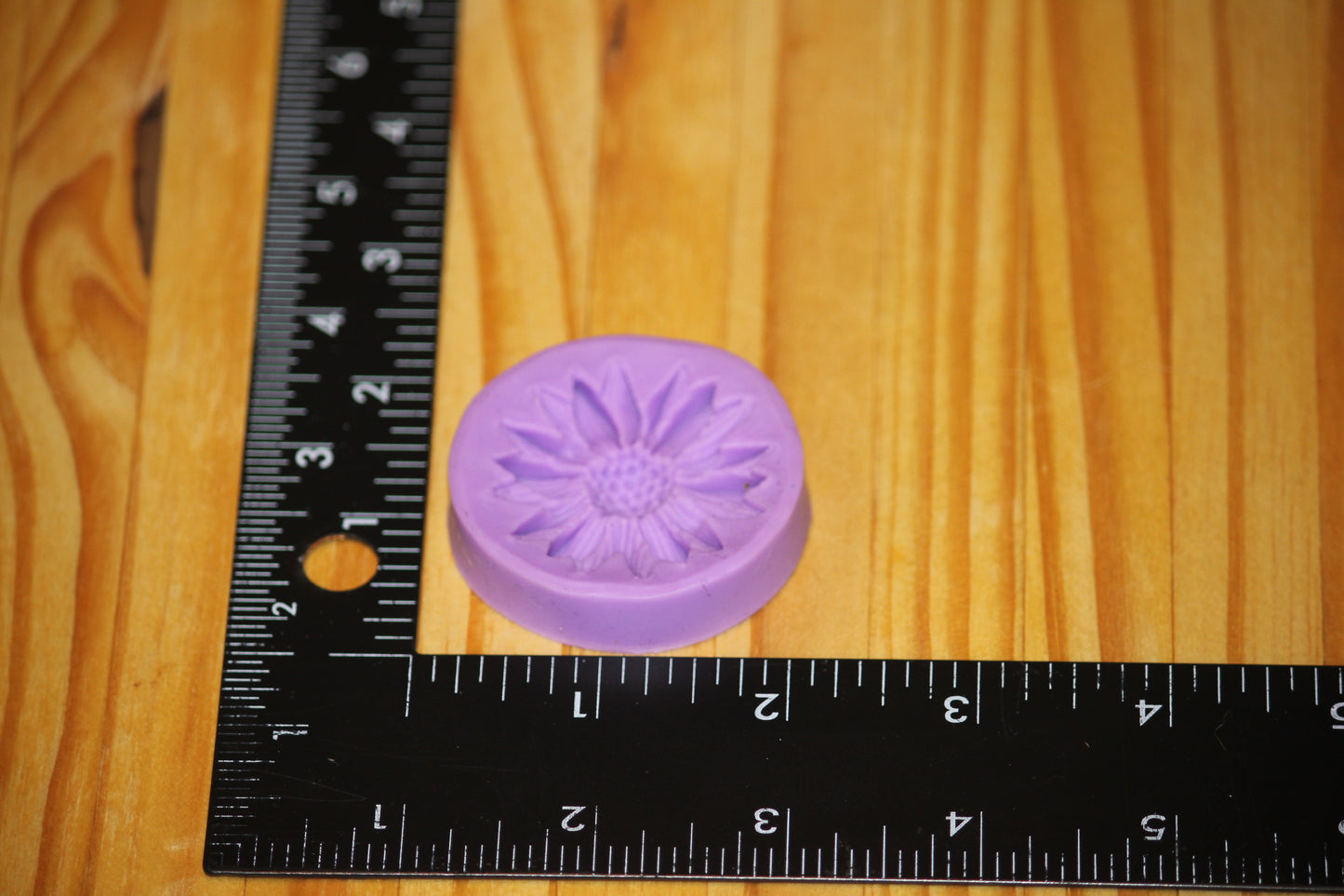 A small lavender purple circular mold with a flower embossed design, shown next to a black ruler on a wooden surface to indicate its size.