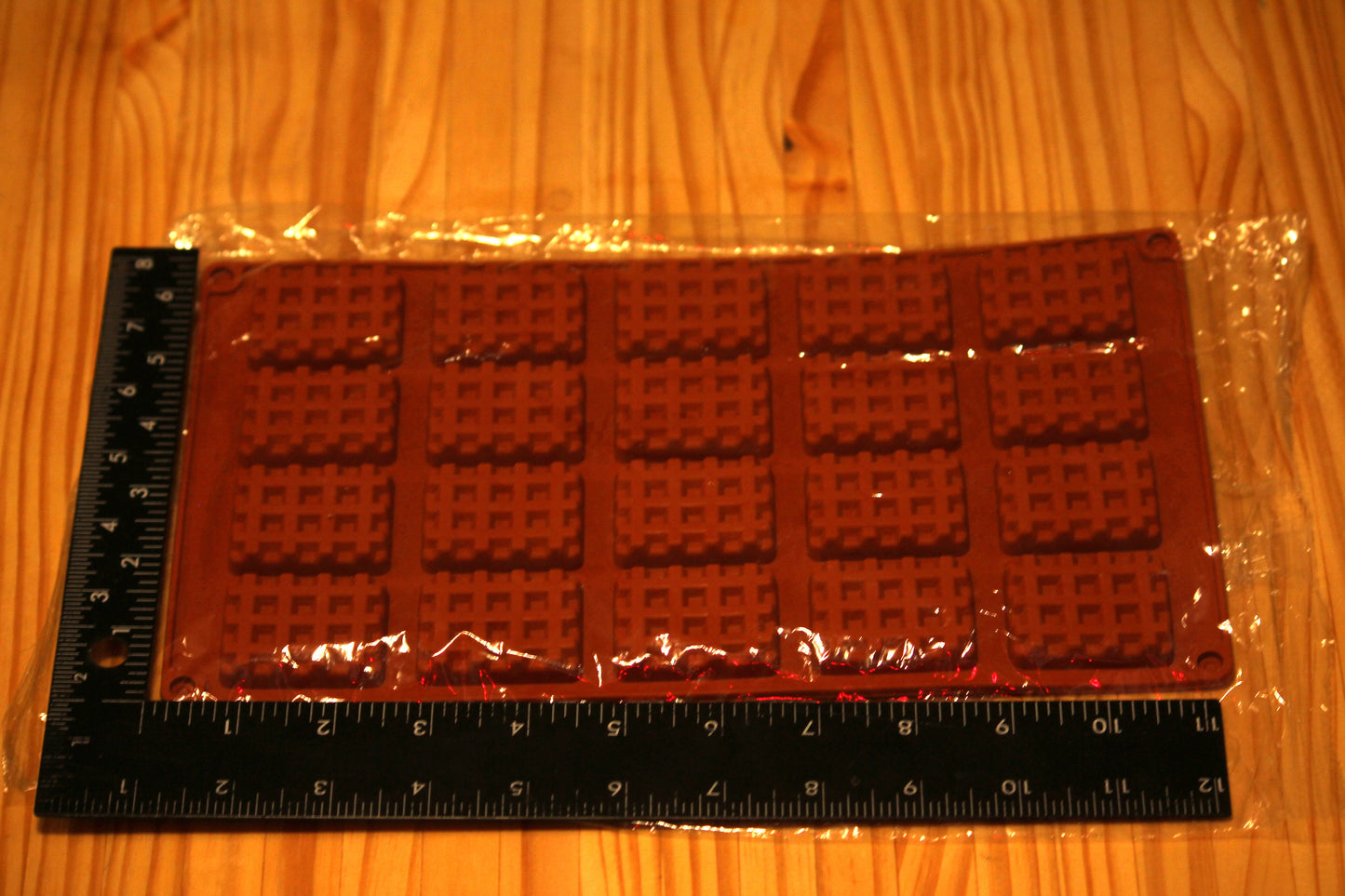 A rectangular chocolate mold or tray with a waffle-like raised pattern, sealed in clear plastic packaging and placed on a wooden surface with black measuring rulers along the left and bottom edges for scale.
