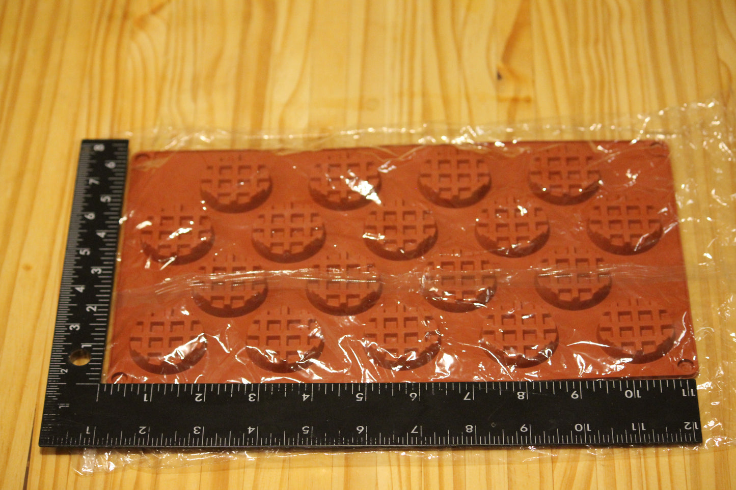 A rectangular brown silicone mold with waffle-shaped cavities, wrapped in clear plastic, shown on a light wood grain surface with a black ruler for scale.