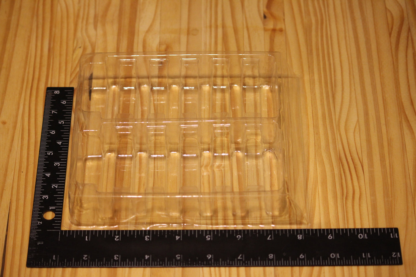 A clear plastic clamshell tray with internal molded inserts, shown on a light wood surface with a black ruler measuring its dimensions.