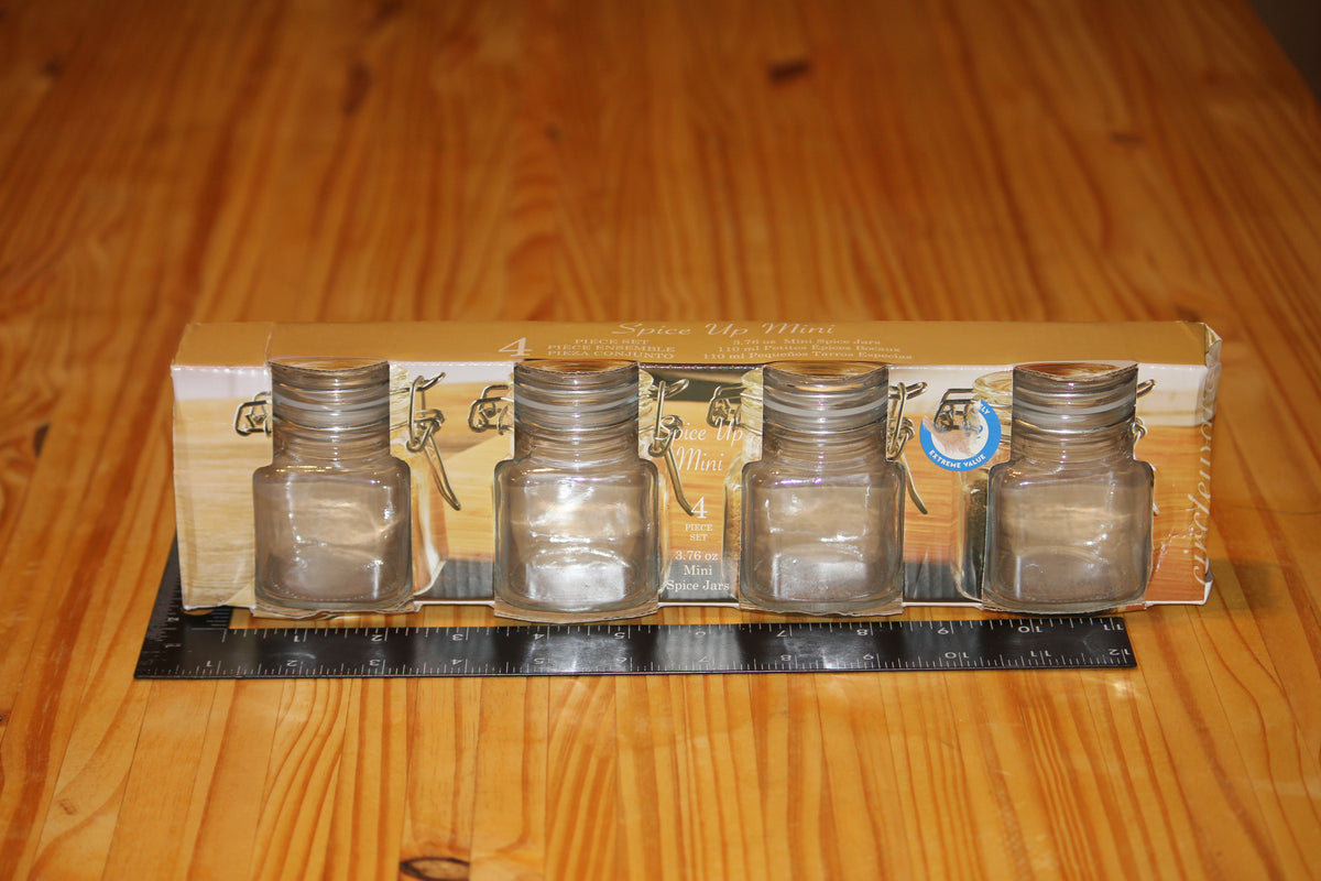 Four small clear glass bottles with clasp caps, shown in their cardboard packaging on a wooden tabletop, with a black ruler placed in front for scale.
