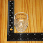 A small clear glass shot glass with a skull shape, shown on a wooden surface next to a black L-shaped measuring ruler to indicate its size.