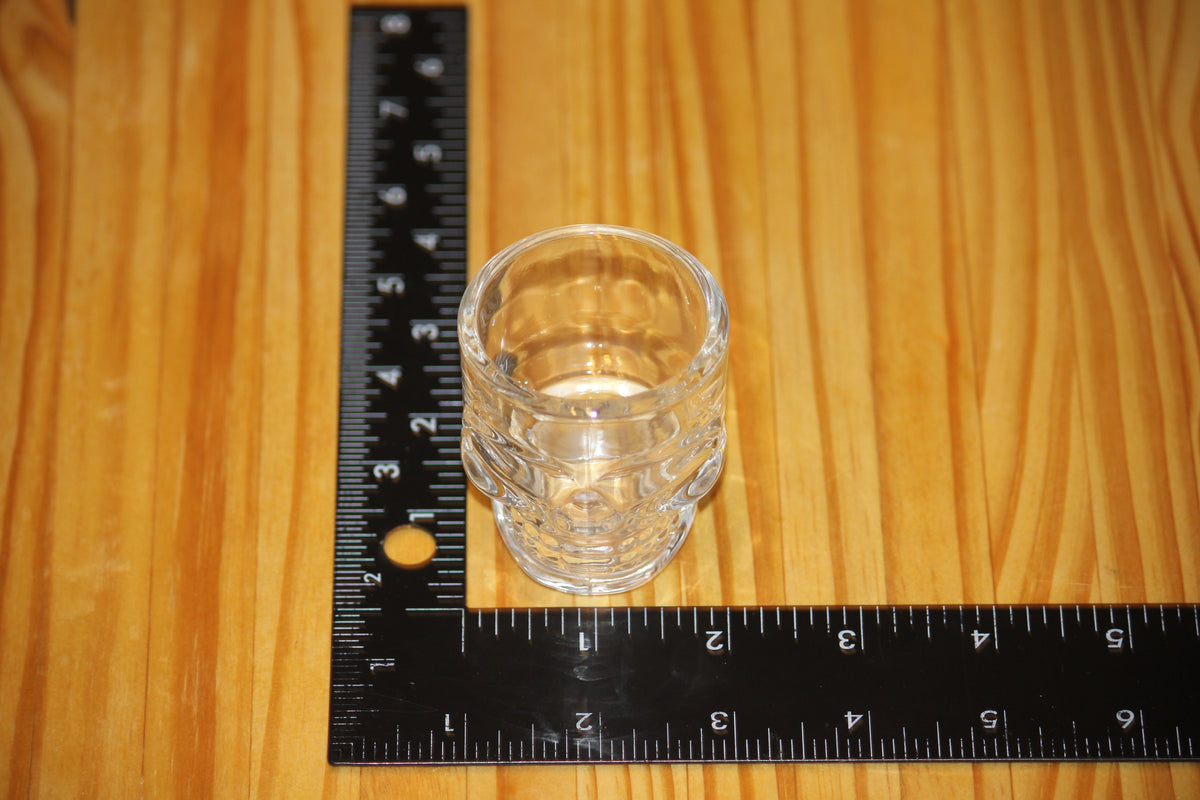 A small clear glass shot glass with a skull shape, shown on a wooden surface next to a black L-shaped measuring ruler to indicate its size.