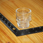 A clear glass skull-shaped shot glass placed on a wooden surface next to a black L-square ruler for scale.