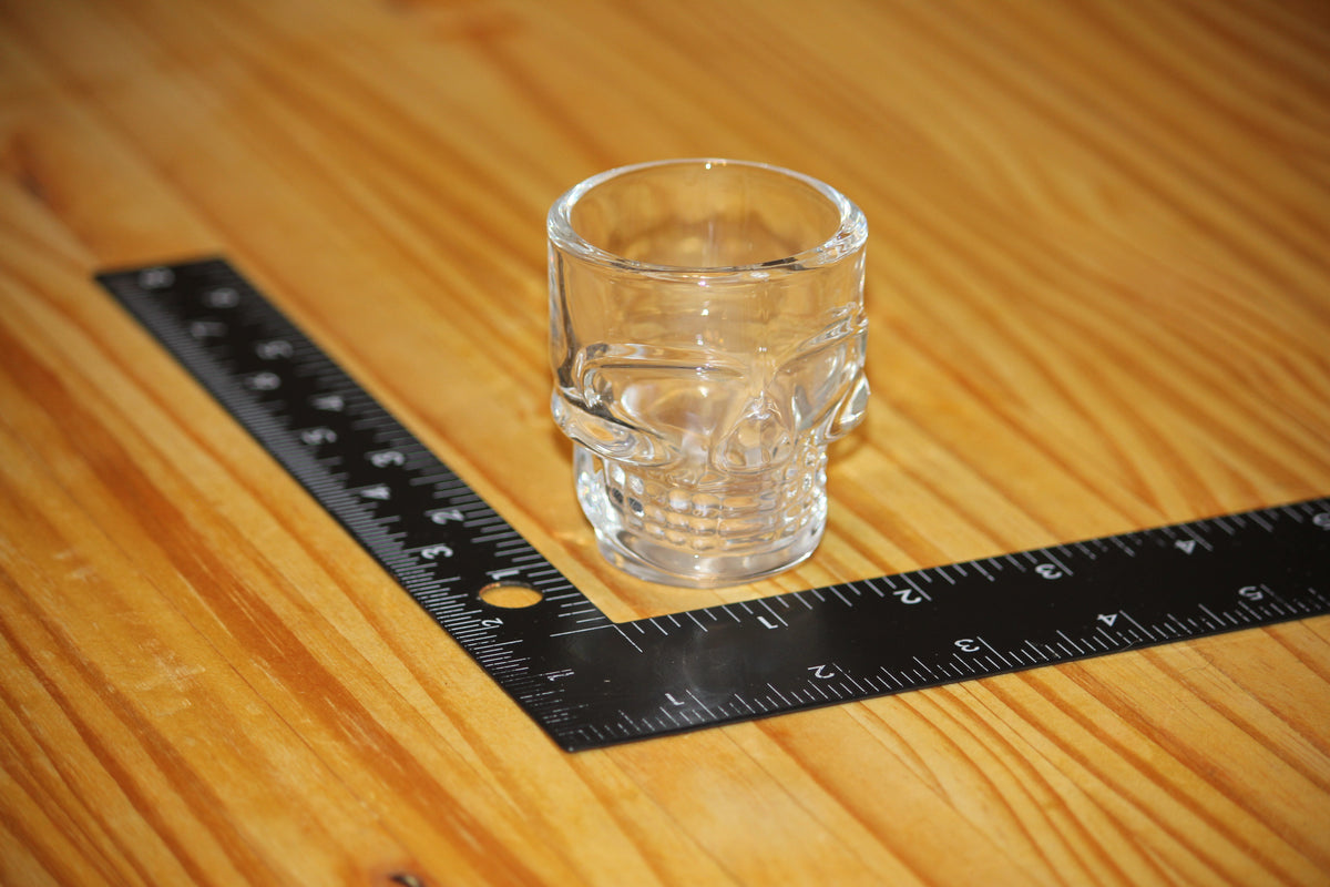 A clear glass skull-shaped shot glass placed on a wooden surface next to a black L-square ruler for scale.