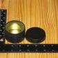 A small round black metal tin with its lid off, placed on a wooden surface next to a black measuring ruler to show size.