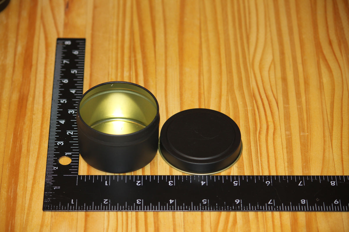 A small round black metal tin with its lid off, placed on a wooden surface next to a black measuring ruler to show size.