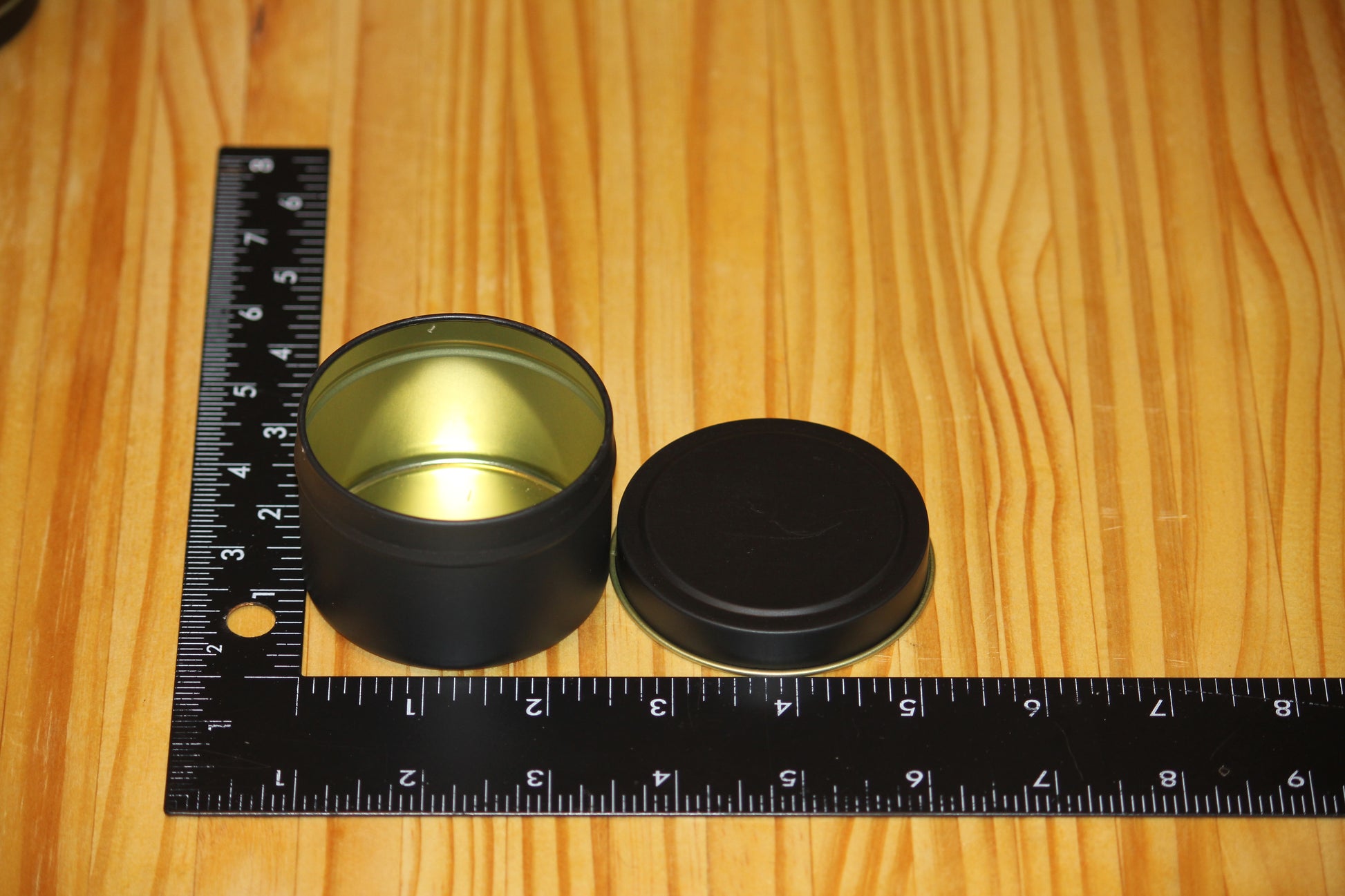 A small round black metal tin with its lid off, placed on a wooden surface next to a black measuring ruler to show size.
