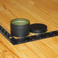 A small round black metal tin with its lid off, placed on a wooden surface next to a black measuring ruler to show size.