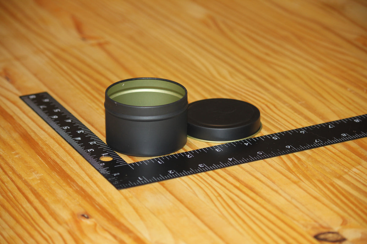 A small round black metal tin with its lid off, placed on a wooden surface next to a black measuring ruler to show size.