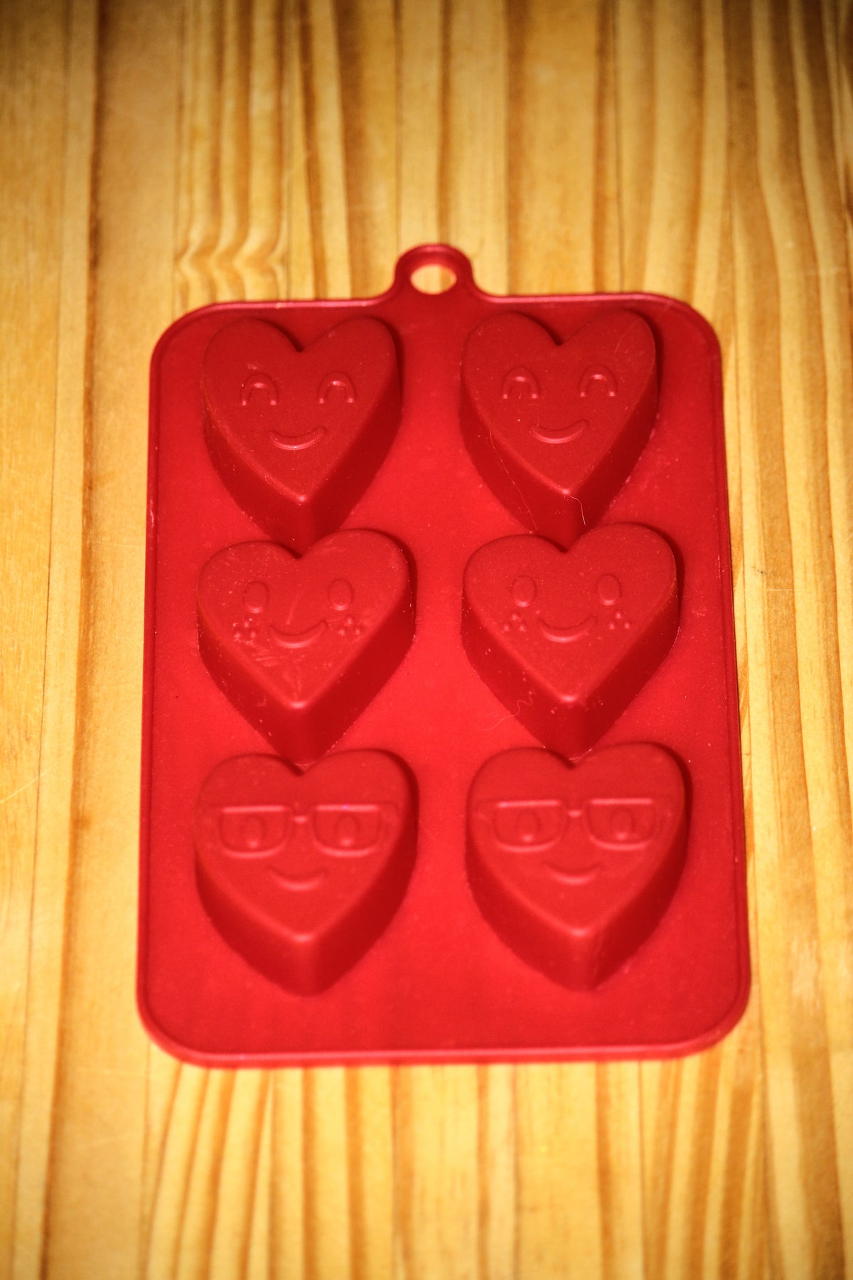 A red silicone mold with six heart-shaped cavities, each featuring a different face design, displayed on a light wood grain surface.