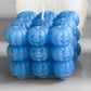 A blue cube-shaped candle made of carved jack-o'-lantern pumpkins, with a white wick, shown on a neutral gray background.