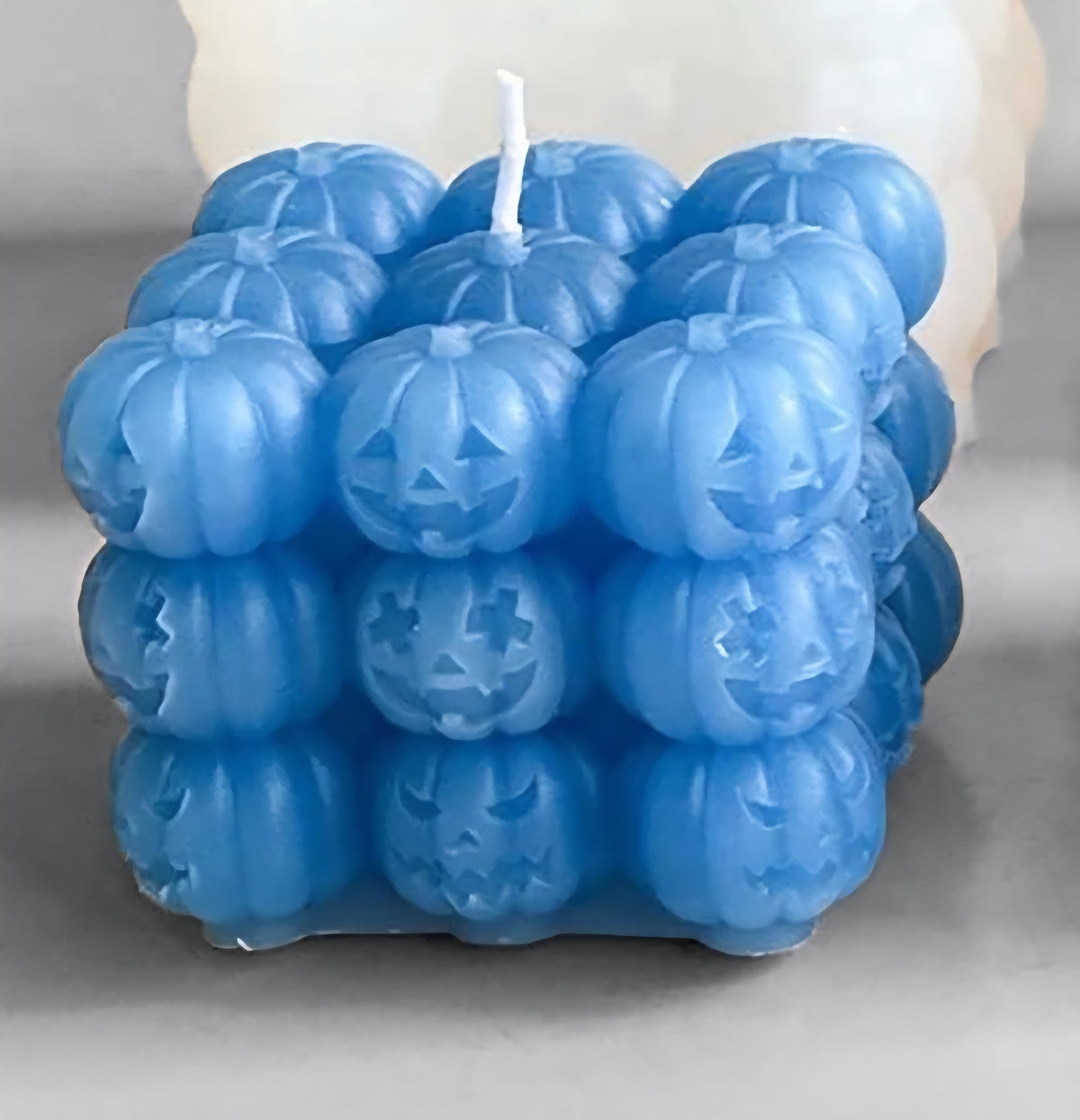 A blue cube-shaped candle made of carved jack-o'-lantern pumpkins, with a white wick, shown on a neutral gray background.
