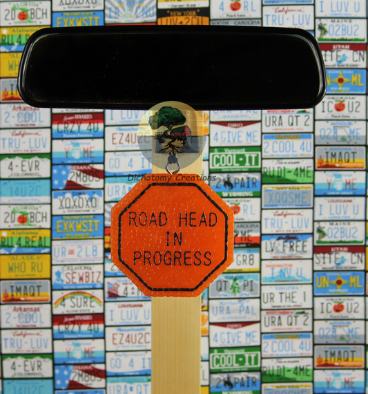 A orange road sign freshie with the words 'ROAD HEAD IN PROGRESS', displayed on a rearview mirror in front of a colorful background of license plates.