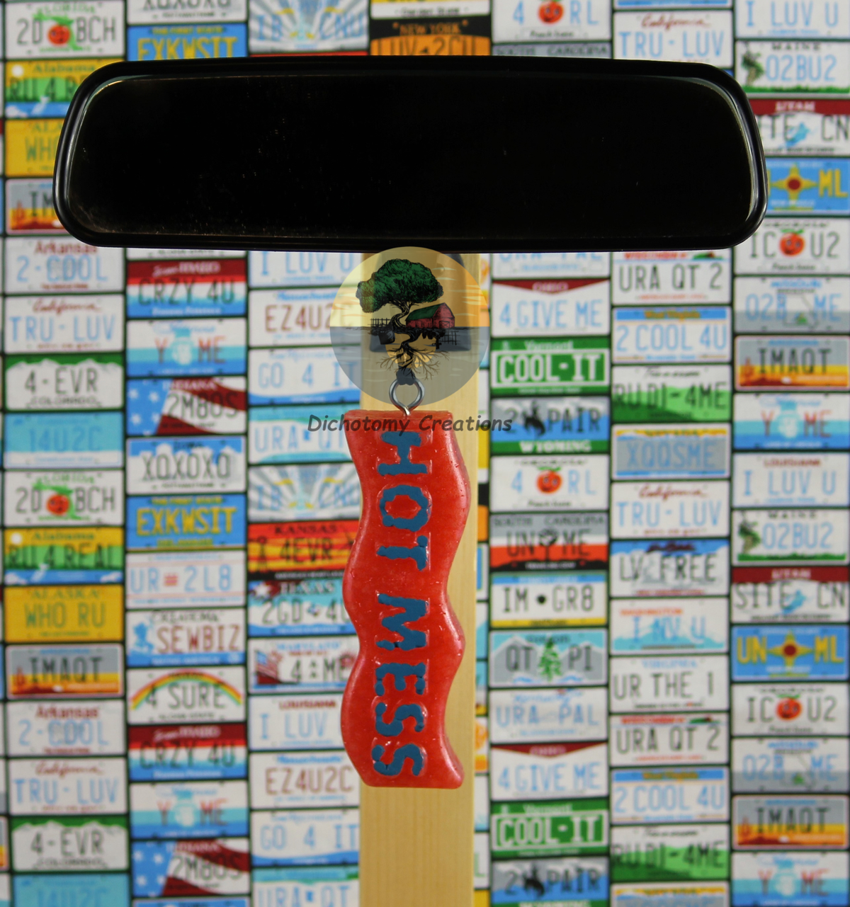 A pink freshie with the words 'HOT MESS' hanging from a rearview mirror against a colorful background of license plates.