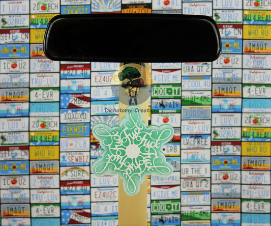 A light blue snowflake freshie, with the word "fuck" hidden in the design.  Displayed from a rearview mirror with a background of colorful license plates.