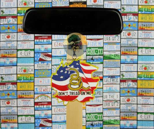 A yellow freshie with a snake and the words 'DON'T TREAD ON ME' with an American flag, displayed on a rearview mirror, set against a background of colorful license plates.