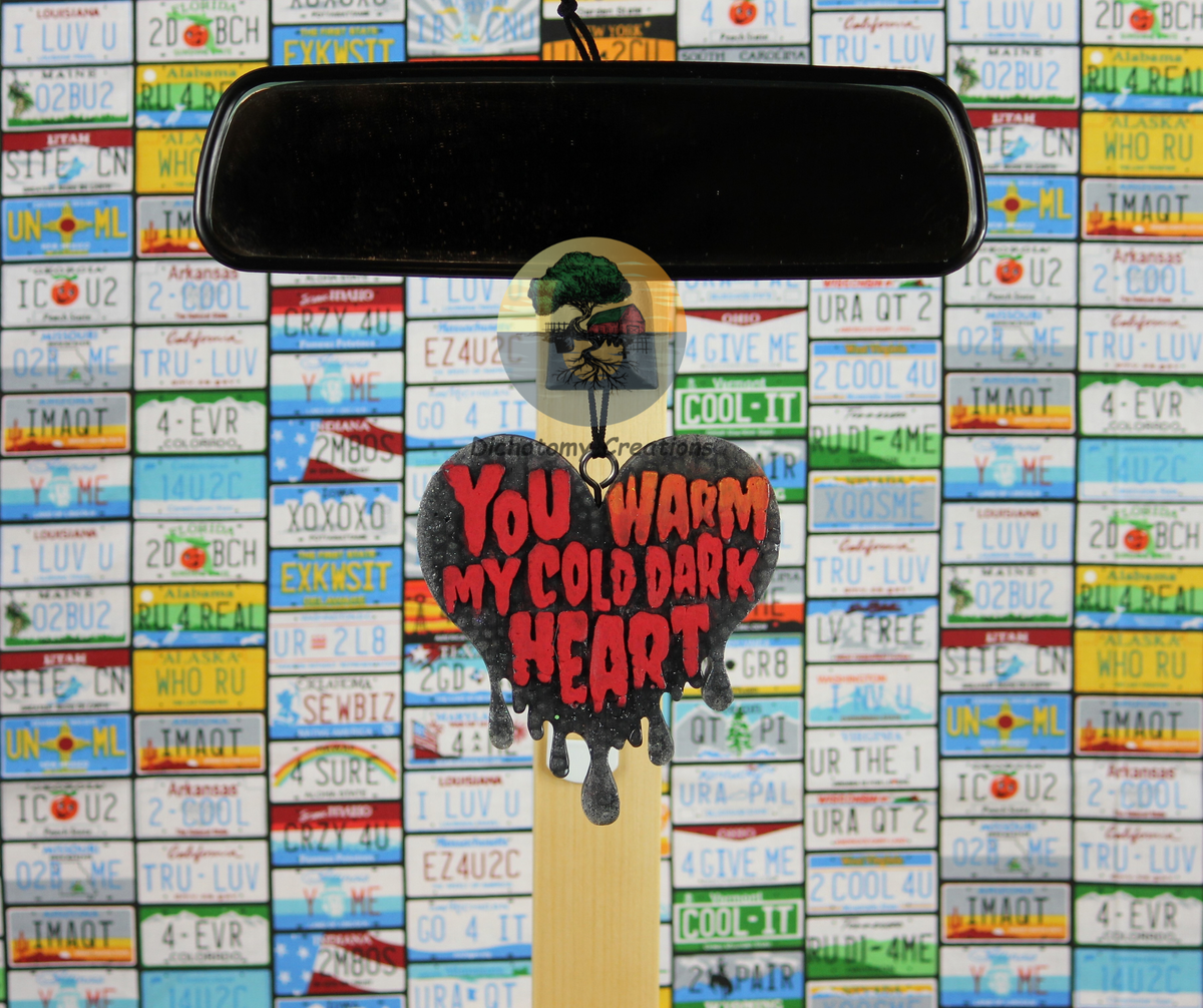 A heart-shaped black freshie with red text reading 'YOU WARM MY COLD DARK HEART' hangs from a rearview mirror. The background is a dense grid of colorful vintage license plates.