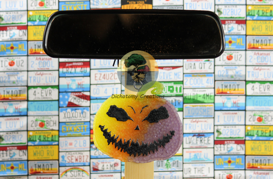 A rainbow Halloween jack-o'-lantern freshie, positioned in front of a rearview mirror. The background is a wall covered in colorful license plates.