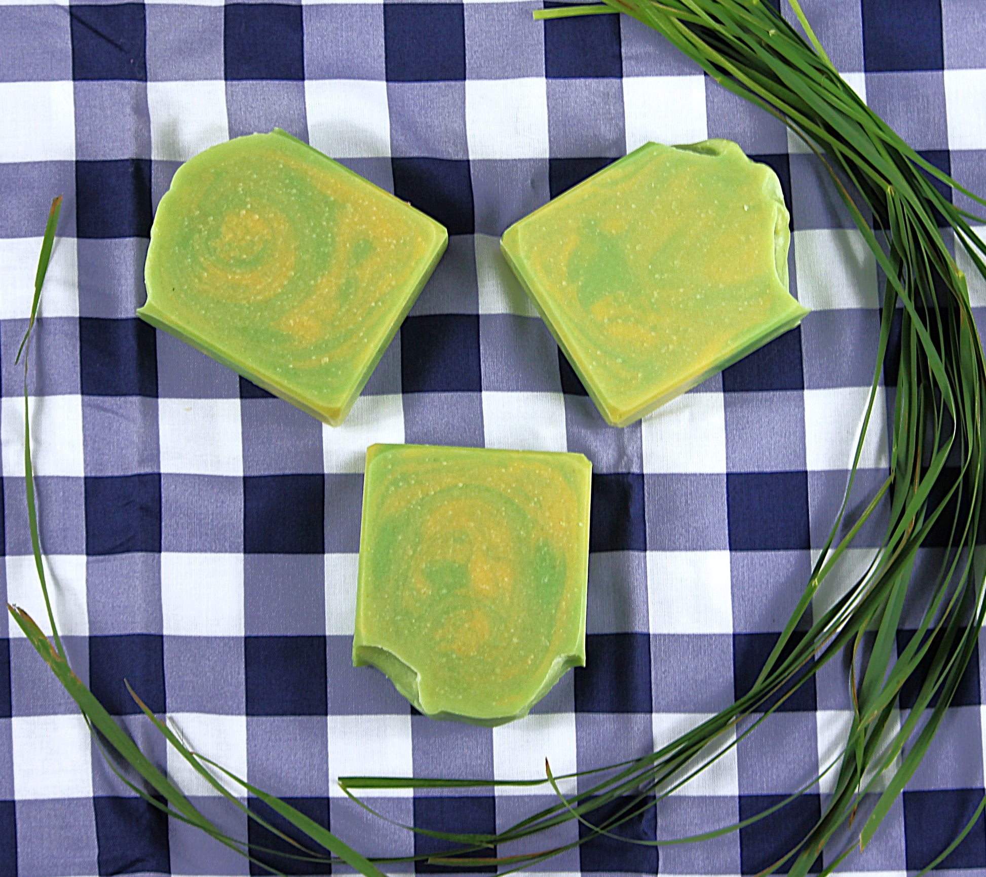 Three green soap bars, encircled by lemongrass, on a blue and white checkered cloth.