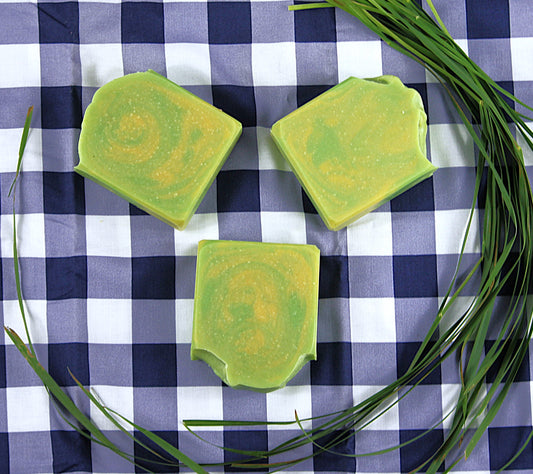 Three green soap bars, encircled by lemongrass, on a blue and white checkered cloth.